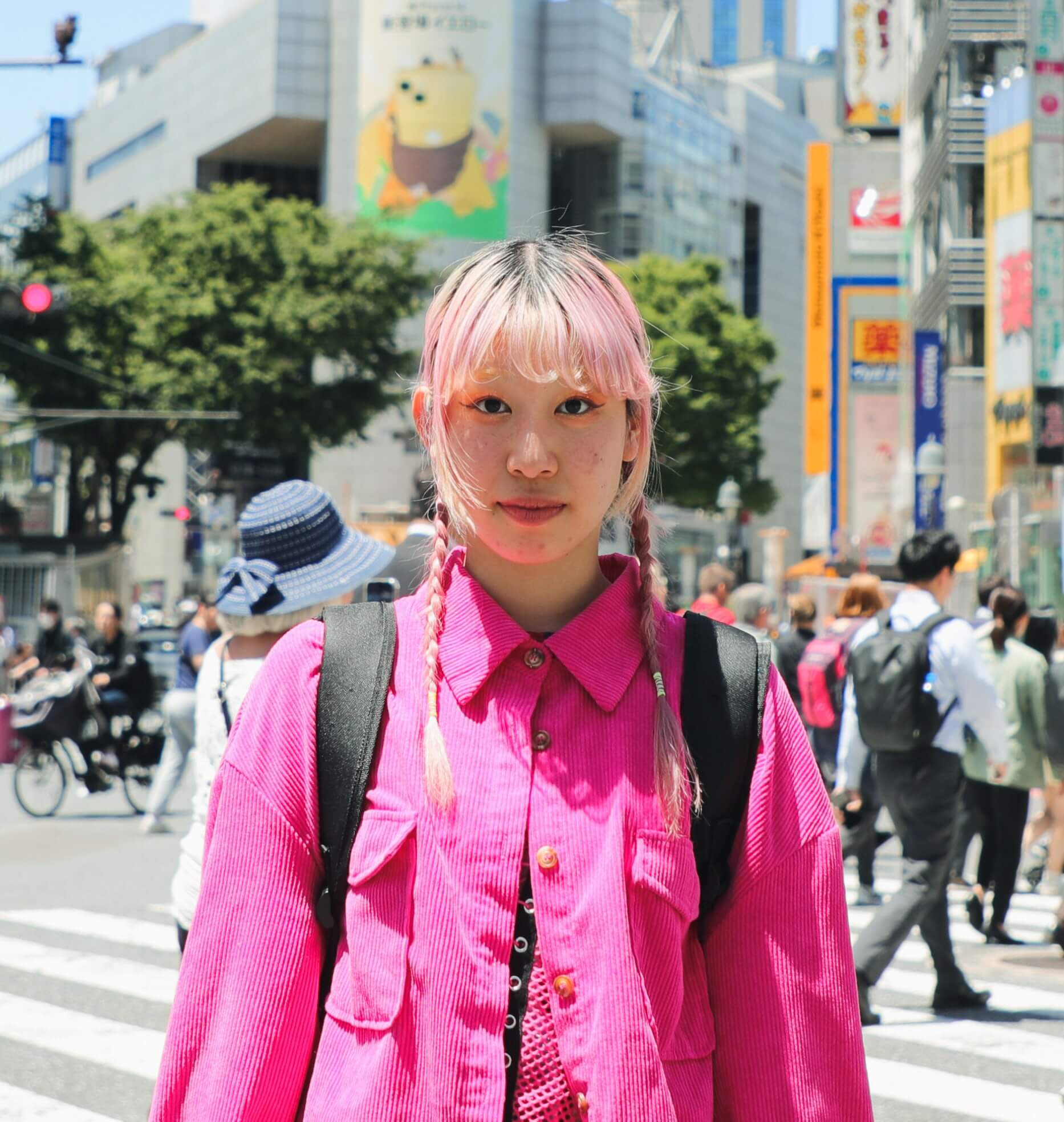 Jovem de cabelo rosa e usando uma camisa rosa está do lado de fora em uma rua movimentada da cidade